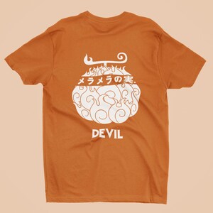 Op de afbeelding: Oranje T-shirt met een witte afbeelding van een gestileerde vrucht met vlammen en het woord "DEVIL" eronder. Japanse tekst is ook zichtbaar. Een uniek T-shirt voor elke garderobe.
