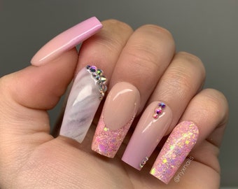 Peut inclure: Ongles longs en acrylique de forme ovale, roses et blancs, avec des paillettes et des strass. Les ongles présentent un motif marbré, des paillettes et des strass. Les ongles sont décorés du texte "@riyxnails".