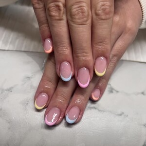 Pode incluir: Close-up de uma mão com unhas ovais. Cada unha apresenta uma ponta francesa em tons pastel de amarelo, rosa, azul e laranja. As unhas têm um acabamento brilhante.
