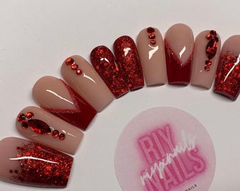 May include: A set of artificial nails in various designs. Some nails are nude with red glitter or a red V-shaped design. Others are solid red glitter. Several nails feature red rhinestones. A pink and white sticker with the text "RIY Nails" is in the image.