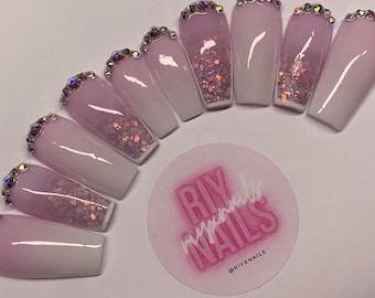 Peut inclure: Des ongles press-on en ombré rose et blanc avec des paillettes et des strass. Les ongles sont longs et de forme de cercueil. Les ongles sont disposés en demi-cercle avec un logo au centre qui dit "RIY press-on NAILS" avec le texte "@RIYXNAILS" sous le logo.