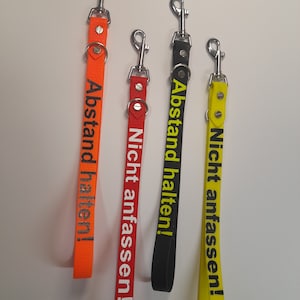 Puede incluir: Tres cordones coloridos con clips metálicos. Los cordones son naranja, negro y amarillo. El texto en los cordones dice "Abstand halten!" y "Nicht anfassen!"
