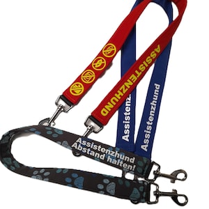 Op de afbeelding: Drie hondenriemen in rood, blauw en zwart met een patroon. De rode riem heeft gele symbolen, de blauwe riem zegt "Assistenzhund", en de zwarte riem "Assistenzhund Abstand halten!" met blauwe pootafdrukken.