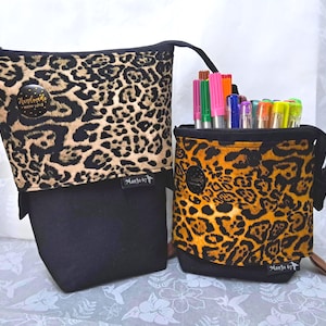 May include: Two fabric pencil cases. One is upright, the other is folded down. Both have a black base and a leopard print pattern in shades of brown and tan. The upright case contains colourful markers. Each case has a small tag that says "ManJo by".
