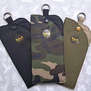 May include: Three eyeglass cases in black, camouflage, and olive green. Each case has a snap closure, a metal ring, and a "Handmade with Love" sticker. The cases are arranged on a white floral patterned surface.