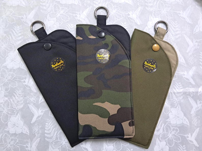 May include: Three eyeglass cases in black, camouflage, and olive green. Each case has a snap closure, a metal ring, and a "Handmade with Love" sticker. The cases are arranged on a white floral patterned surface.