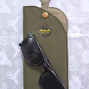 May include: Black sunglasses rest on a green fabric case with a gold snap closure and a metal ring. A small, round sticker with the words "Handmade with Love" is affixed to the case. The background is a white floral pattern.