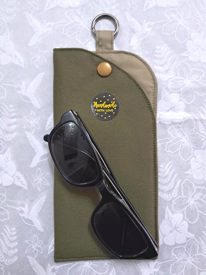 May include: Black sunglasses rest on a green fabric case with a gold snap closure and a metal ring. A small, round sticker with the words "Handmade with Love" is affixed to the case. The background is a white floral pattern.