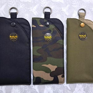 May include: Three rectangular fabric cases with key rings. One is black, one is camouflage, and the third is olive green. Each case has a snap closure and a circular sticker that reads "Handmade with Love."