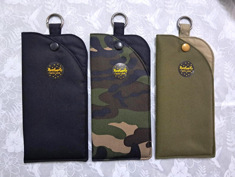 May include: Three rectangular fabric cases with key rings. One is black, one is camouflage, and the third is olive green. Each case has a snap closure and a circular sticker that reads "Handmade with Love."