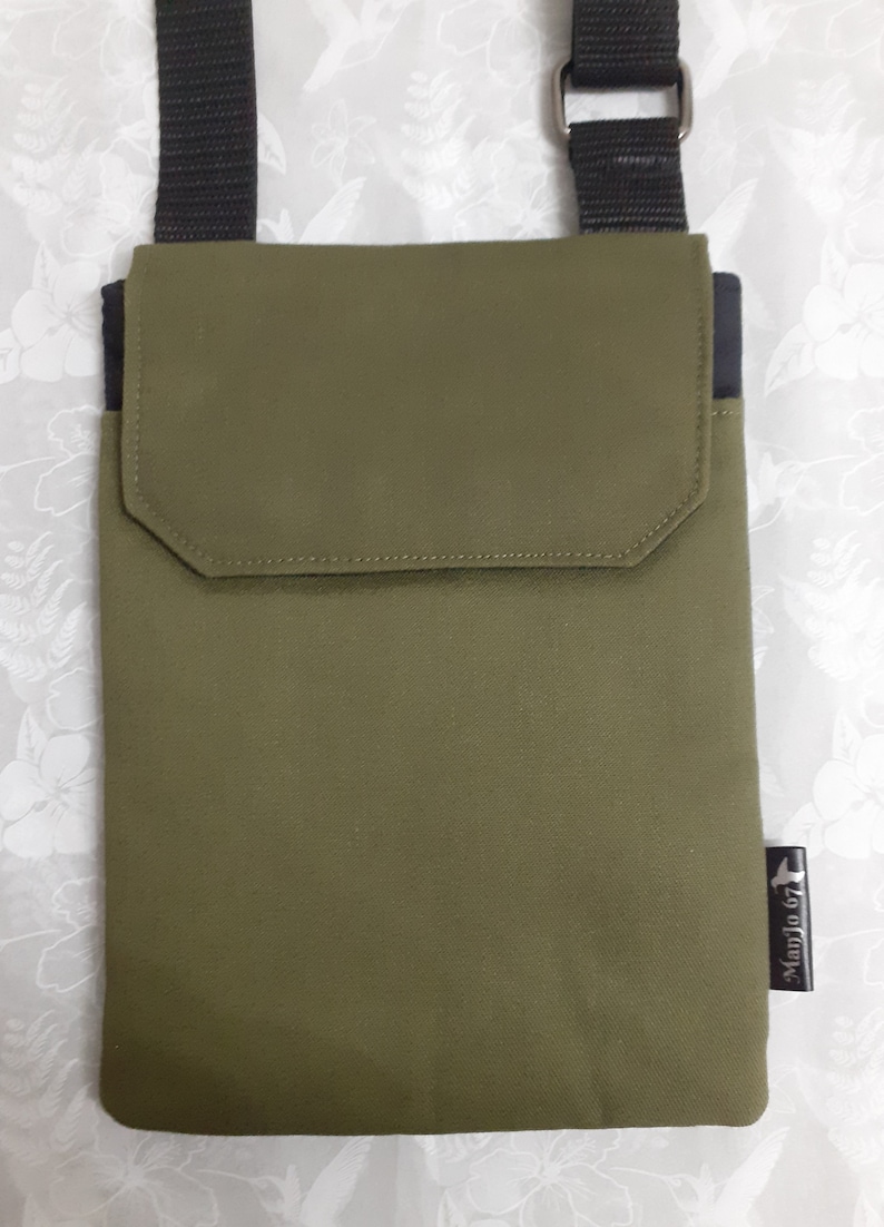 May include: A green canvas crossbody bag with a black strap and a black pocket on the front. The bag has a small black label with the text "Madjo 67" on the bottom right corner.