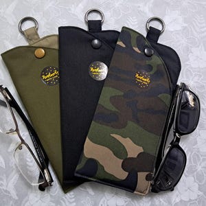 May include: Three eyeglass cases in olive green, black, and camouflage patterns, each with a metal ring and snap closure. One case holds eyeglasses, and another holds sunglasses. Each case has a sticker that says "Handmade with love."