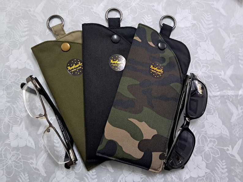 May include: Three eyeglass cases in olive green, black, and camouflage patterns, each with a metal ring and snap closure. One case holds eyeglasses, and another holds sunglasses. Each case has a sticker that says "Handmade with love."