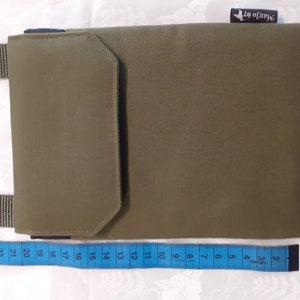 May include: A green canvas crossbody bag with a flap closure and a smaller pocket on the front. The bag has a black strap and a blue label with the text "Manjo 67" on the top right corner. The bag is measured with a blue tape measure.