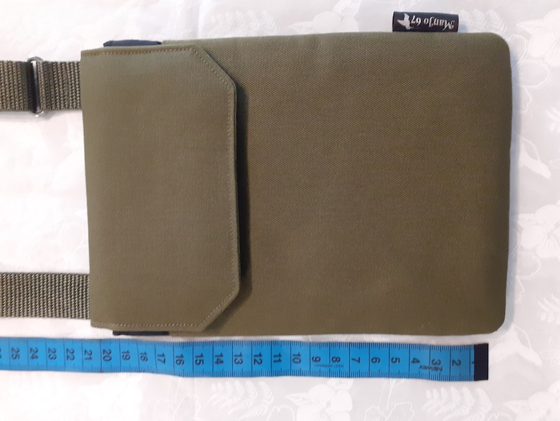 May include: A green canvas crossbody bag with a flap closure and a smaller pocket on the front. The bag has a black strap and a blue label with the text "Manjo 67" on the top right corner. The bag is measured with a blue tape measure.