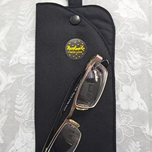 May include: Black eyeglass case with a silver metal ring and snap closure. The case holds a pair of eyeglasses with silver frames. A yellow sticker with the words "Handmade with Love" is on the case.