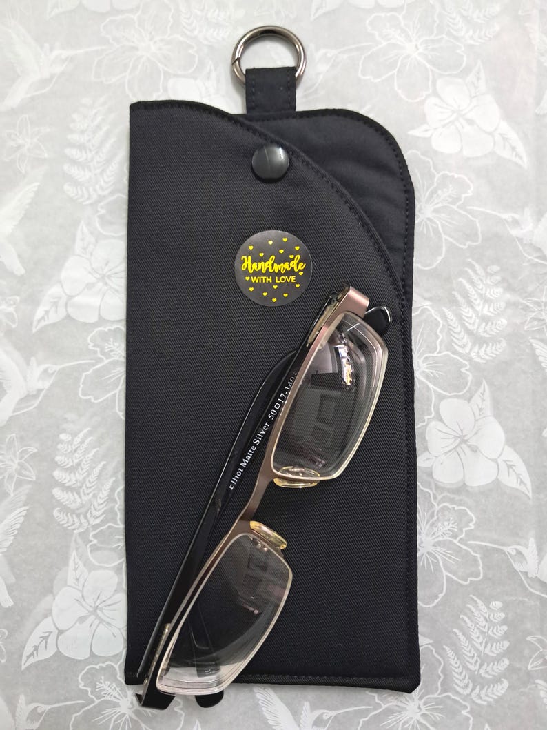 May include: Black eyeglass case with a silver metal ring and snap closure. The case holds a pair of eyeglasses with silver frames. A yellow sticker with the words "Handmade with Love" is on the case.