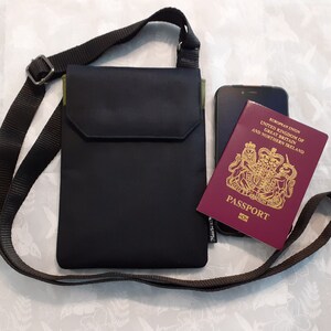 May include: A black crossbody bag with a zippered pocket and a green accent. The bag is lying on a white surface with a passport and a cell phone inside.