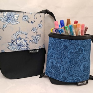 May include: Two fabric pencil cases. One is grey with blue floral patterns and a black base. The other is blue with a paisley design and a black base. Both have black zips and handles. The blue case is filled with colourful pens and markers.