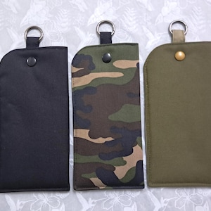 May include: Three key fob holders in black, camouflage, and olive green. Each holder has a metal ring at the top and a snap closure. The camouflage pattern features shades of green, brown, and black.