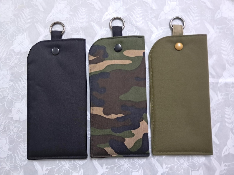 May include: Three key fob holders in black, camouflage, and olive green. Each holder has a metal ring at the top and a snap closure. The camouflage pattern features shades of green, brown, and black.