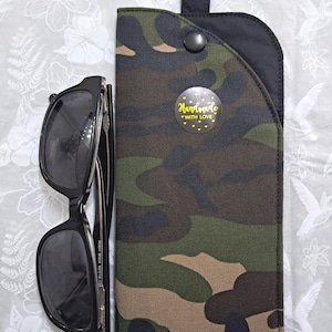 May include: Black sunglasses next to a camouflage print glasses case. The case has a black top with a metal ring and a snap closure. A sticker on the case reads "Handmade with Love" with yellow hearts.