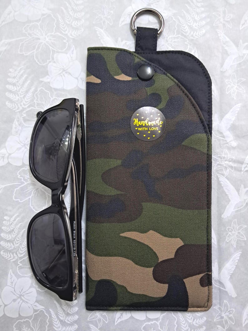 May include: Black sunglasses next to a camouflage print glasses case. The case has a black top with a metal ring and a snap closure. A sticker on the case reads "Handmade with Love" with yellow hearts.