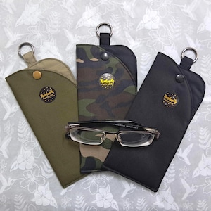 May include: Three eyeglass cases in olive green, camouflage, and black, each with a snap closure and a metal ring. A pair of eyeglasses rests on the camouflage case. Each case has a small, round sticker that says "Handmade with Love."