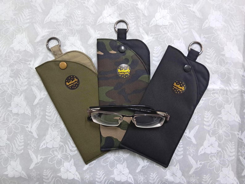 May include: Three eyeglass cases in olive green, camouflage, and black, each with a snap closure and a metal ring. A pair of eyeglasses rests on the camouflage case. Each case has a small, round sticker that says "Handmade with Love."