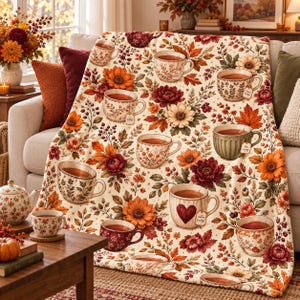 May include: A cream-colored throw blanket featuring a repeating pattern of various tea cups, some with tea tags labeled 'tea' or 'cozy', interspersed with vibrant autumn-themed illustrations of orange, red, and yellow flowers and fall leaves.