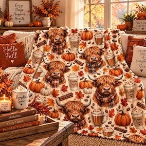May include: A cream-coloured throw blanket featuring a repeating pattern of Highland cows wearing plaid scarves, orange pumpkins, cinnamon sticks, and cups of pumpkin spice lattes with whipped cream, accented by scattered autumn leaves.