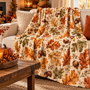 May include: A cream-colored throw blanket featuring a repeating pattern of autumn oak leaves in shades of orange, brown, and muted green, interspersed with detailed illustrations of brown acorns throughout the fabric.