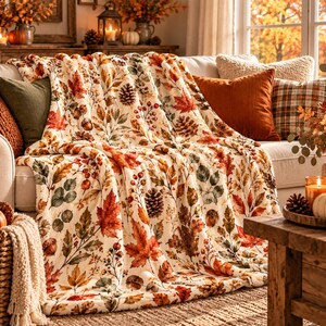 May include: A cream-colored throw blanket featuring a repeating pattern of autumn leaves in shades of orange, red, and brown, interspersed with illustrations of pinecones and acorns. The fabric has a soft, plush texture draped over a sofa.