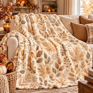 May include: A cream-colored plush throw blanket featuring a repeating pattern of autumn leaves and berries in shades of brown, tan, and muted orange. The soft, textured fabric is draped over a sofa, showcasing detailed botanical illustrations.