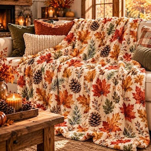 May include: A cream-colored throw blanket featuring a repeating pattern of vibrant orange, red, and yellow maple leaves, green pine needles, and brown pinecones, draped over a sofa with several decorative pillows.
