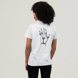 May include: White t-shirt with a black graphic of a hand on the back. The hand illustration features intricate details, including lines and star-like shapes. The shirt is a classic crew neck style.