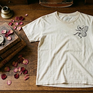 May include: Cream-colored t-shirt featuring a black illustration of a fairy with butterfly wings. The tee has a crew neck and short sleeves. The shirt is displayed on a wooden surface with antique books, a compass, and rose petals.