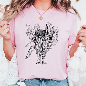 May include: A light pink t-shirt featuring a black and white botanical illustration of a plant with large leaves and a central flower. The design is centered on the shirt. The shirt is being worn.