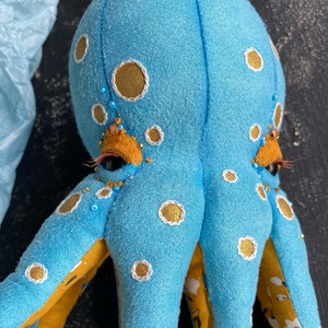 May include: A blue octopus plush toy with yellow and white polka dots and gold accents. The octopus has black eyes and a beaded fringe around the eyes.