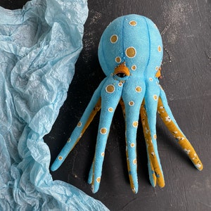 May include: A blue and yellow octopus plush toy with white polka dots. The octopus has eight tentacles and is sitting on a blue tissue paper background.