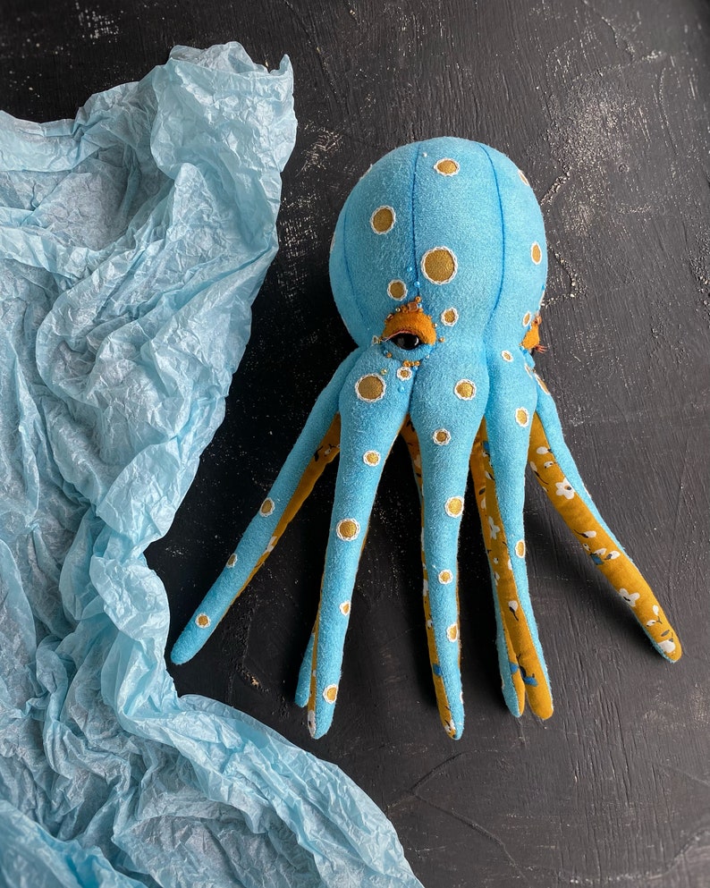May include: A blue and yellow octopus plush toy with white polka dots. The octopus has eight tentacles and is sitting on a blue tissue paper background.