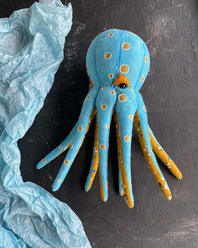 May include: A blue and yellow octopus plush toy with white polka dots. The octopus has eight tentacles and a large, round head with a single eye.