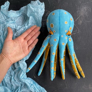May include: A blue and yellow octopus plush toy with white polka dots and embroidered eyes. The octopus is lying on a blue tissue paper background.