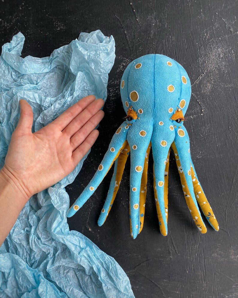 May include: A blue and yellow octopus plush toy with white polka dots and embroidered eyes. The octopus is lying on a blue tissue paper background.