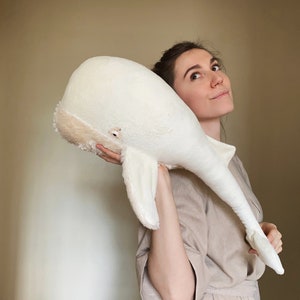 Puede incluir: Una mujer sostiene un gran juguete de peluche de ballena blanca frente a ella. La ballena es suave y esponjosa con una nariz marr&oacute;n claro y un peque&ntilde;o ojo negro.