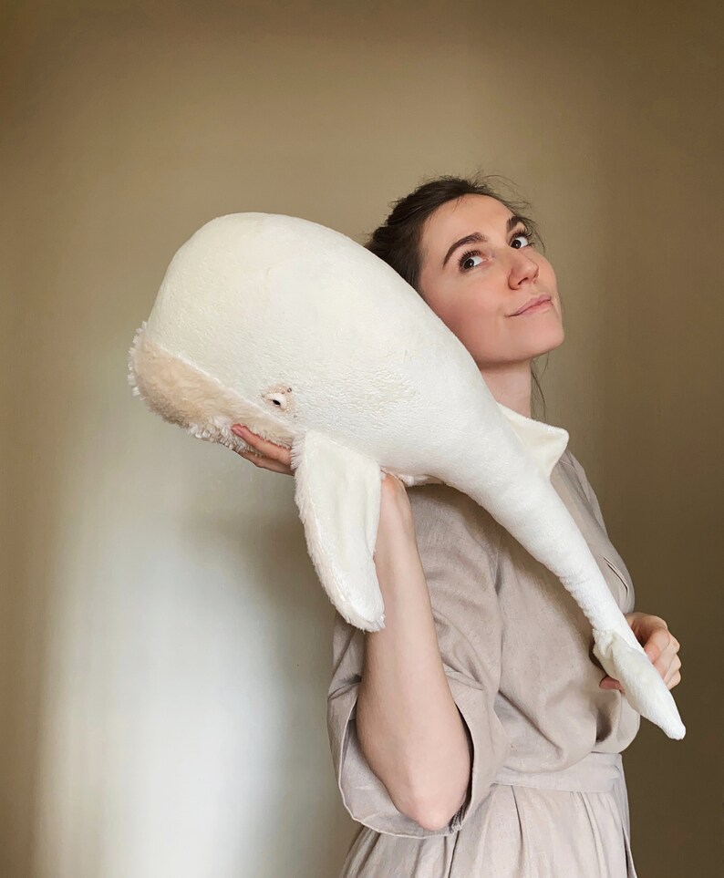 Puede incluir: Una mujer sostiene un gran juguete de peluche de ballena blanca frente a ella. La ballena es suave y esponjosa con una nariz marr&oacute;n claro y un peque&ntilde;o ojo negro.