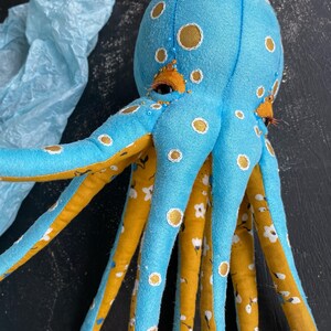 May include: A blue octopus plush toy with yellow and white accents. The octopus has eight tentacles and is decorated with white polka dots and a floral pattern.