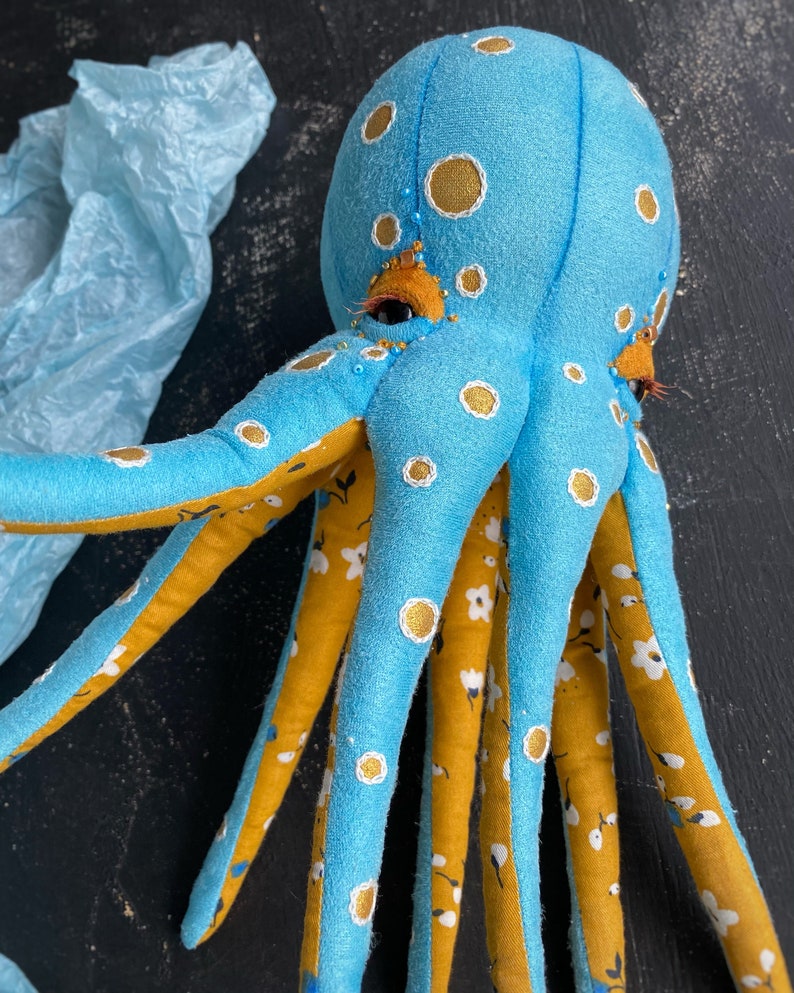 May include: A blue octopus plush toy with yellow and white accents. The octopus has eight tentacles and is decorated with white polka dots and a floral pattern.