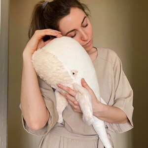 Puede incluir: Una mujer con un vestido beige sostiene un gran juguete de peluche de ballena blanca. Est&aacute; abrazando la ballena y tiene los ojos cerrados.