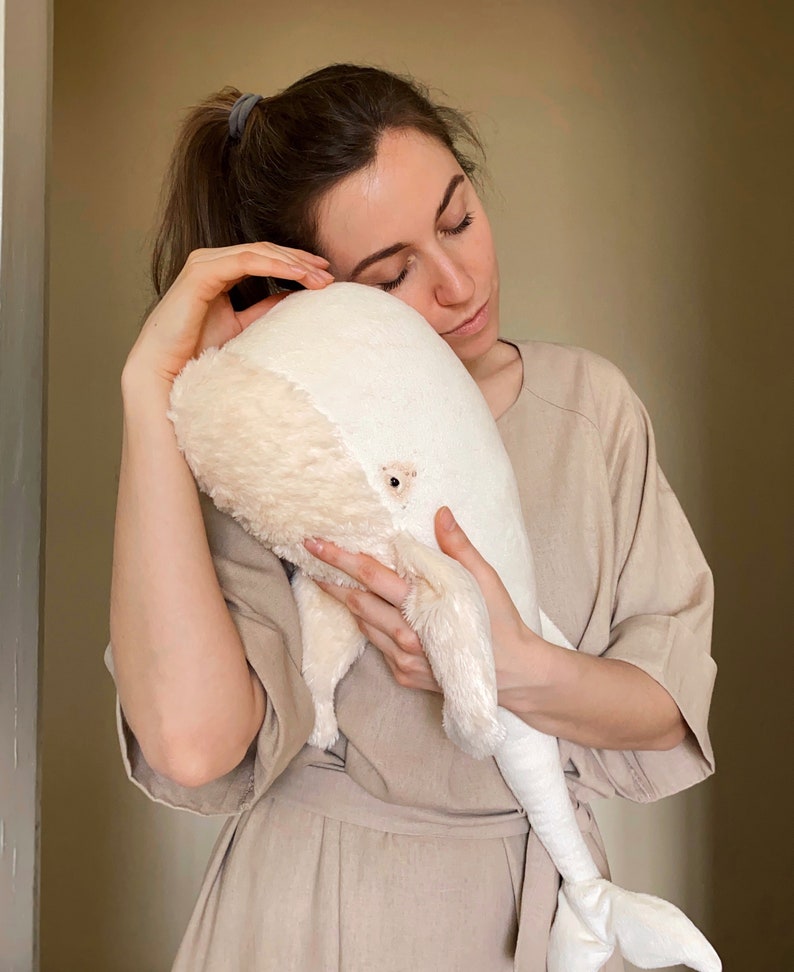 Puede incluir: Una mujer con un vestido beige sostiene un gran juguete de peluche de ballena blanca. Est&aacute; abrazando la ballena y tiene los ojos cerrados.
